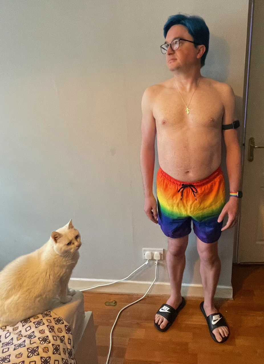 Me in Pride swimming shorts, looking to the side, as a cat watches.